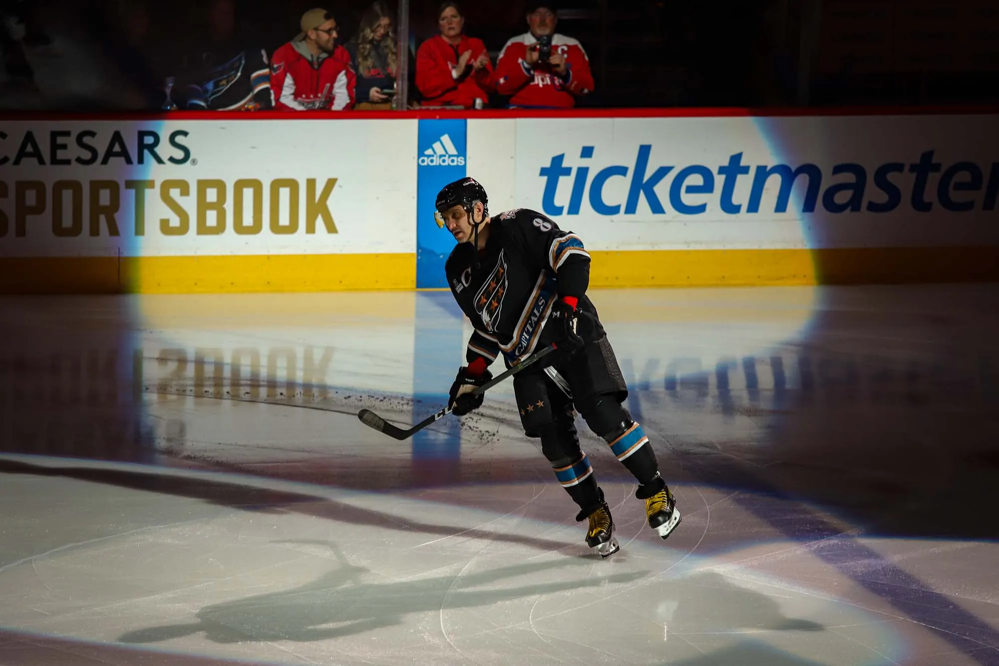 A photo of a hockey player, Alexander Ovechkin, wearing a mostly black uniform skates casually under a spotlight before the start of a game.