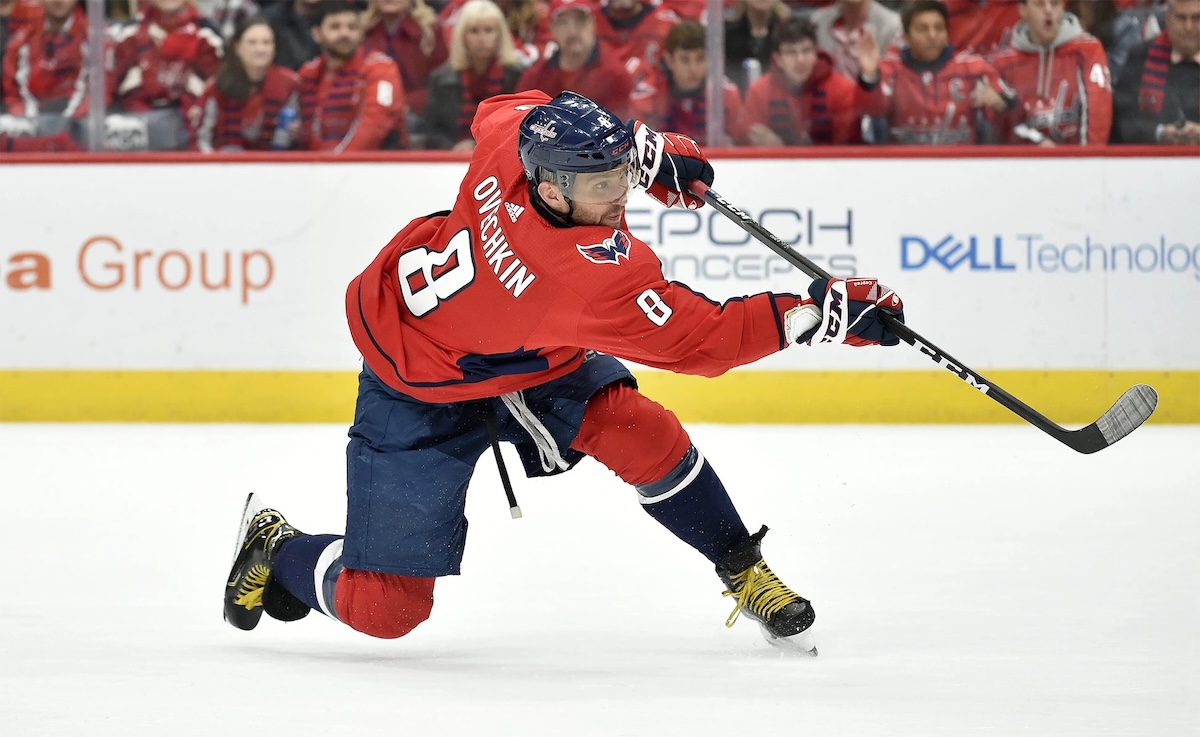 An action shot of a hockey player, Alexander Ovechkin #8 (in a red uniform), in the middle of hitting the puck to the right with his stick outstretched.