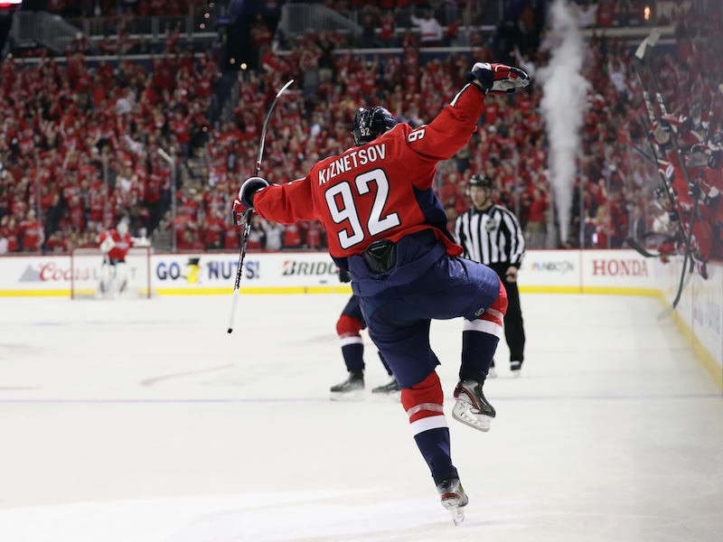 A photo of #92, Evgeny Kuznetsov, celebrating a goal by skating down the ice with both arms and a leg raised to imitate a bird.