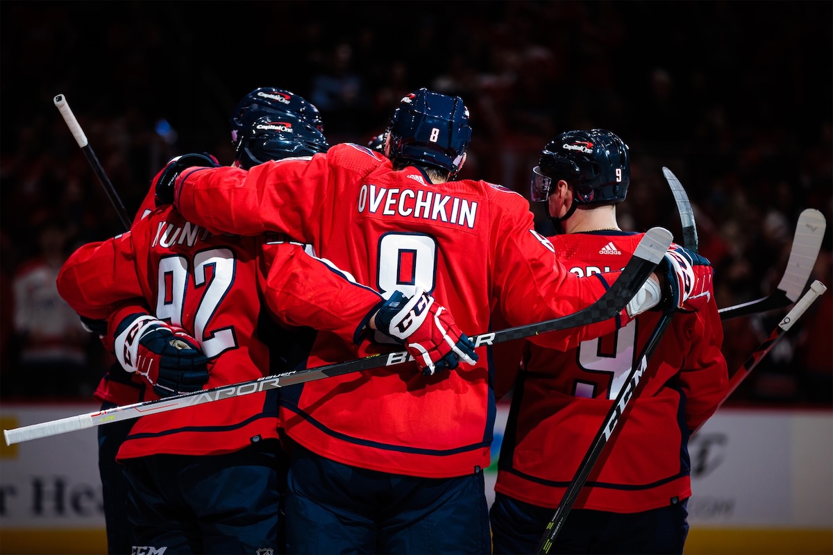 A photo of 4 hockey players in their red uniforms huddled together on the ice as they celebrate a goal.