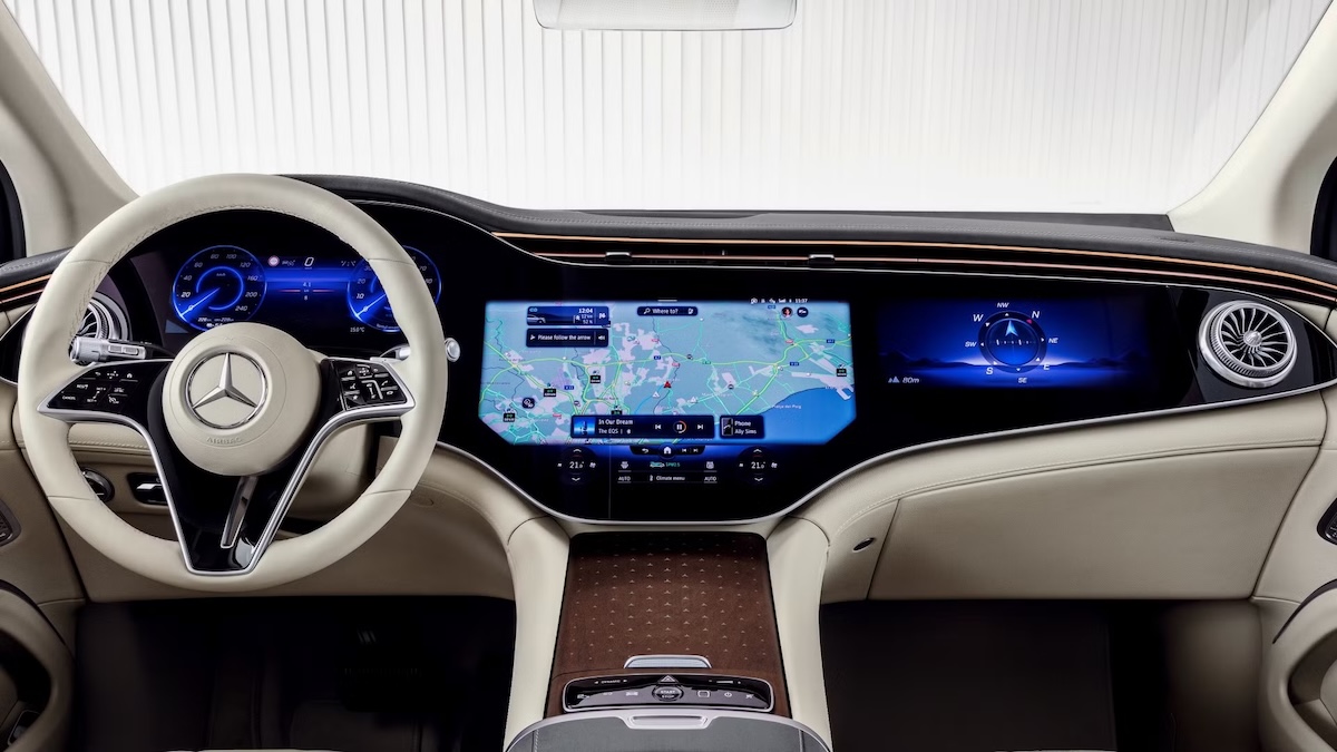 A photo of the interior of an upcoming 2025 Mercedes Benz EQS, showing the entire dashboard. The entire dashboard has been consolidated into 3 touchscreens connected by glossy plastic trying to make it appear like one, large touchscreen.